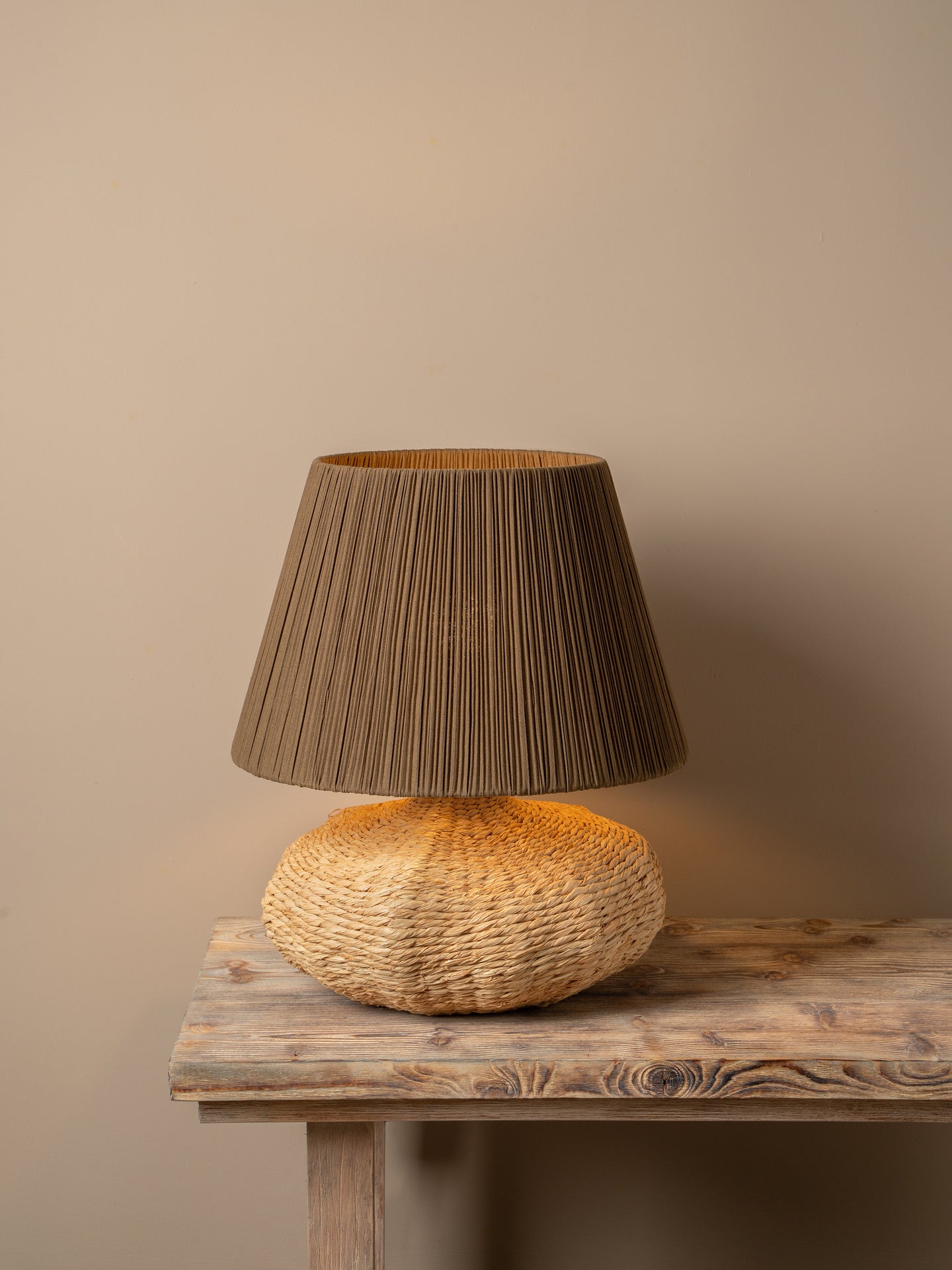 A table lamp with a dark brown pleated fabric shade and a textured natural woven base, illuminated and placed on a rustic wooden table against a warm neutral wall.