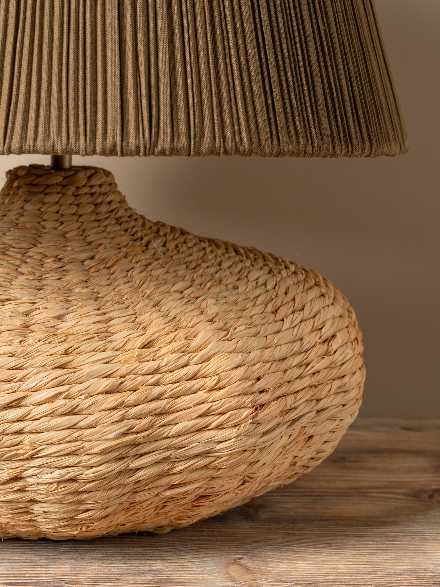 close up of a table lamp with a dark brown pleated fabric shade and a textured natural woven base, placed on a rustic wooden table against a warm neutral wall.