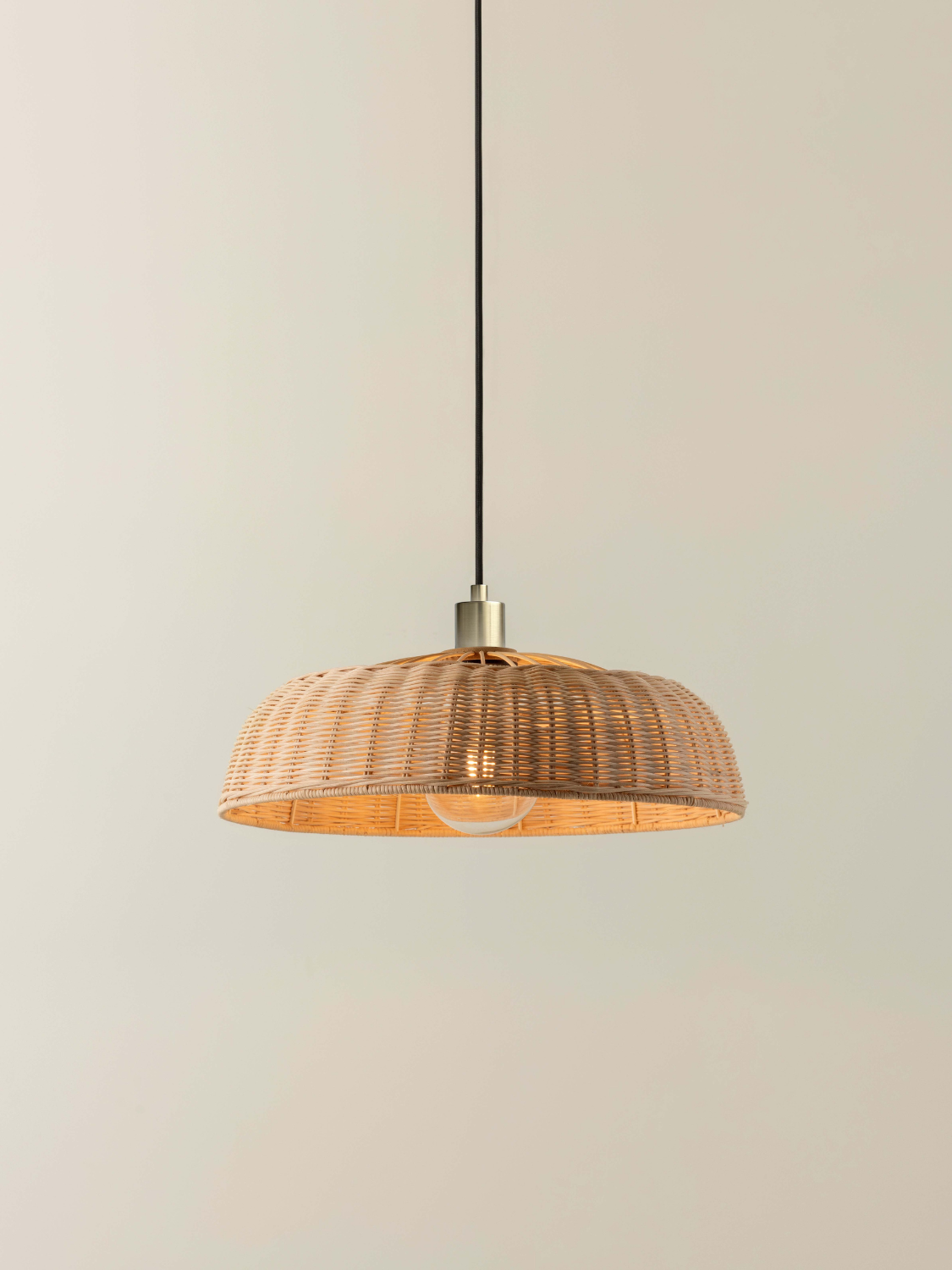 A woven natural rattan pendant light shade with a warm golden-brown finish, suspended by a black cord with a brass metal cap against a neutral beige background.