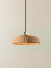 A woven natural rattan pendant light shade with a warm golden-brown finish, suspended by a black cord with a brass metal cap against a neutral beige background.