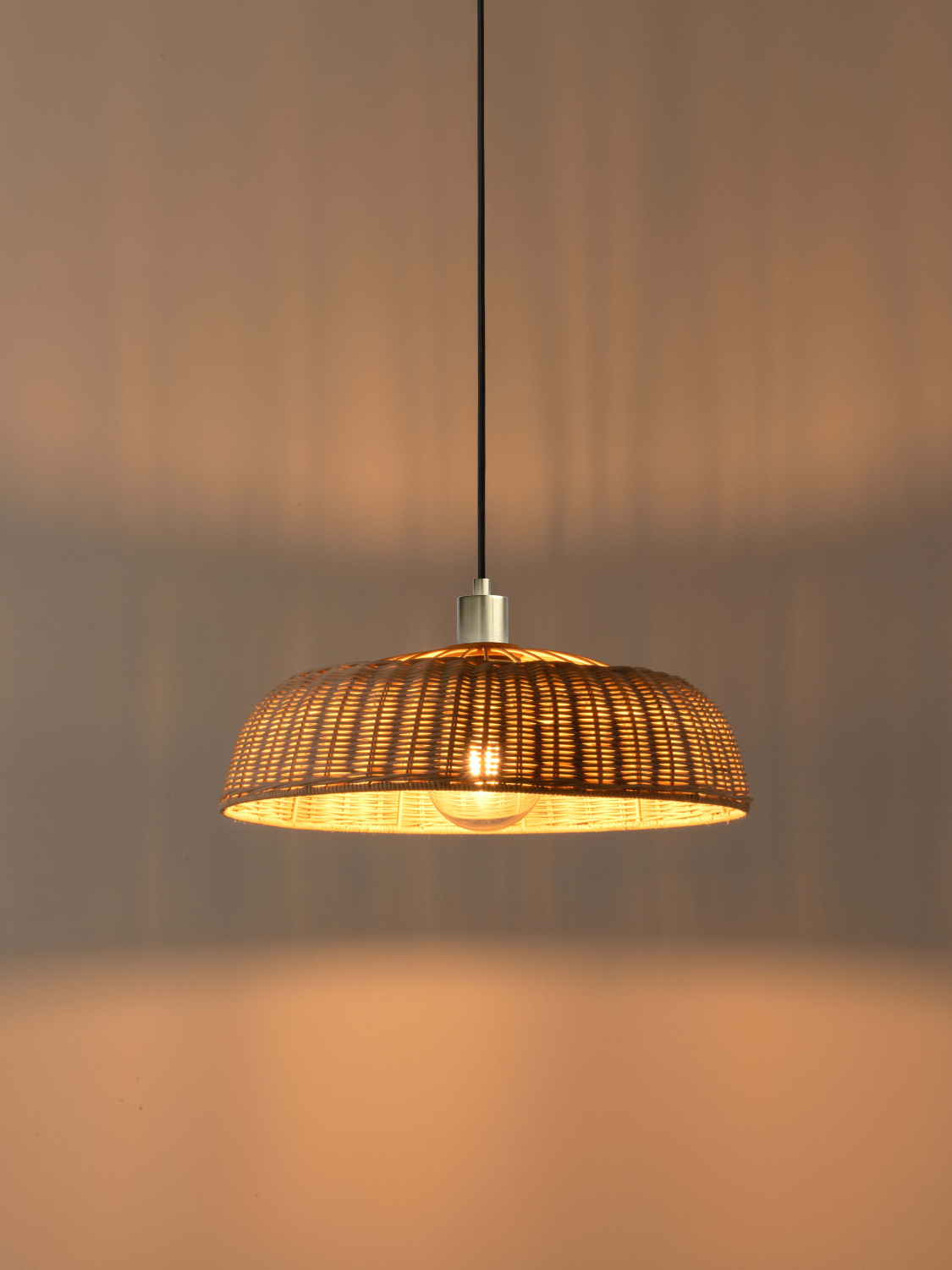 A woven rattan pendant light with a warm glowing bulb inside, suspended by a black cord against a softly lit beige wall.
