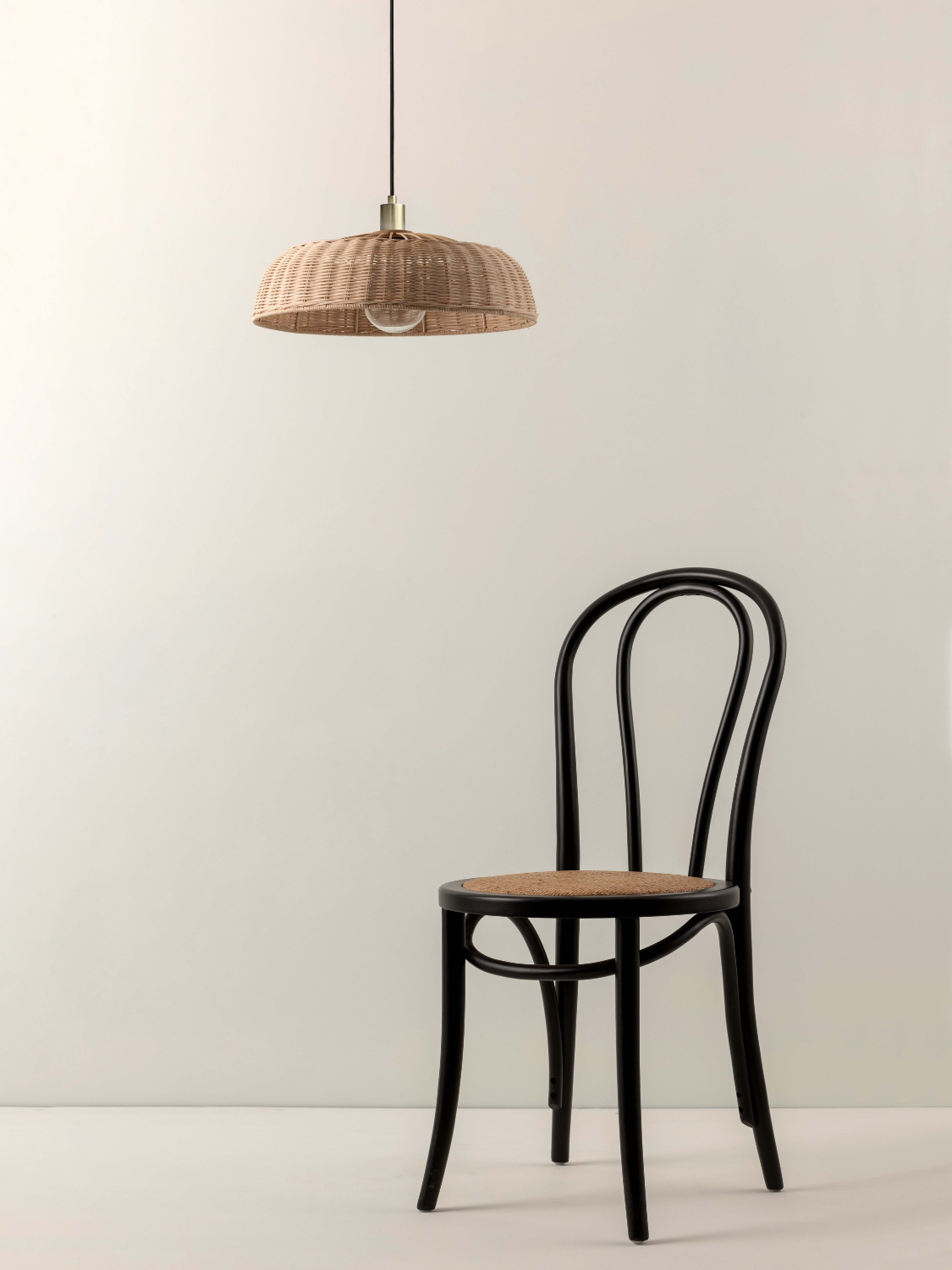A black bentwood chair with a light brown woven seat, positioned below a matching woven pendant lamp shade against a plain off-white wall.