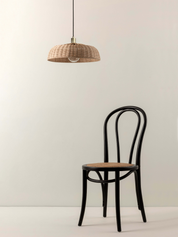 A black bentwood chair with a light brown woven seat, positioned below a matching woven pendant lamp shade against a plain off-white wall.