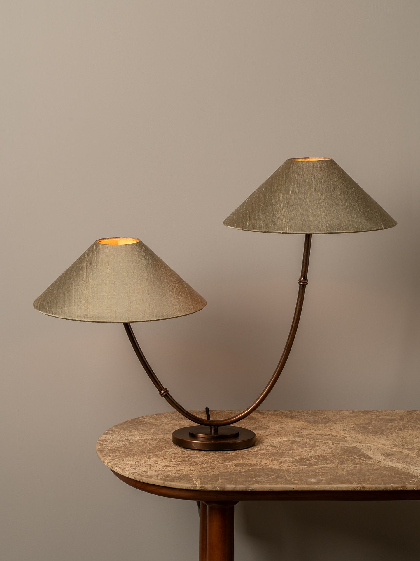A two-light table lamp with conical beige fabric shades and a curved bronze metal base, shown lit on a stone-topped table against a neutral gray wall.