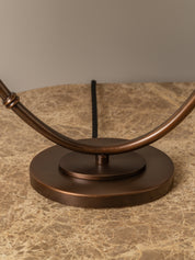 Close-up of a bronze metal table lamp base and curved arm on a beige marble surface against a neutral gray background.