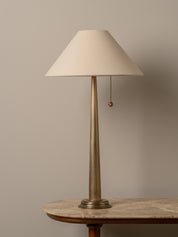 A table lamp with a tapered off-white fabric shade, a slim brushed brass metal stem, and a pull chain, displayed on a stone-topped side table against a neutral beige wall.