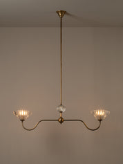 A two-light brass chandelier with curved arms, clear ribbed glass shades, and a central clear glass sphere, shown hanging against a plain beige wall.