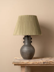 A table lamp with a dark brown ceramic vase-shaped base and a pleated olive green fabric shade, illuminated with a warm glow on a rustic wooden table against a neutral beige wall.