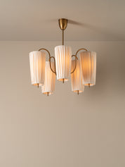A modern five-light chandelier with pleated off-white cylindrical shades and a brushed brass metal frame, suspended from a matching ceiling canopy against a neutral beige wall.