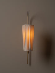A modern wall sconce with a vertical pleated white shade and a slim brass fixture, glowing with a warm orange light against a dark taupe wall.