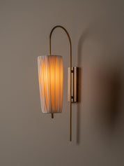 A modern wall-mounted sconce with a warm glowing cylindrical pleated shade, a curved brass-toned metal arm, and a rectangular wall mount, shown against a plain taupe wall.