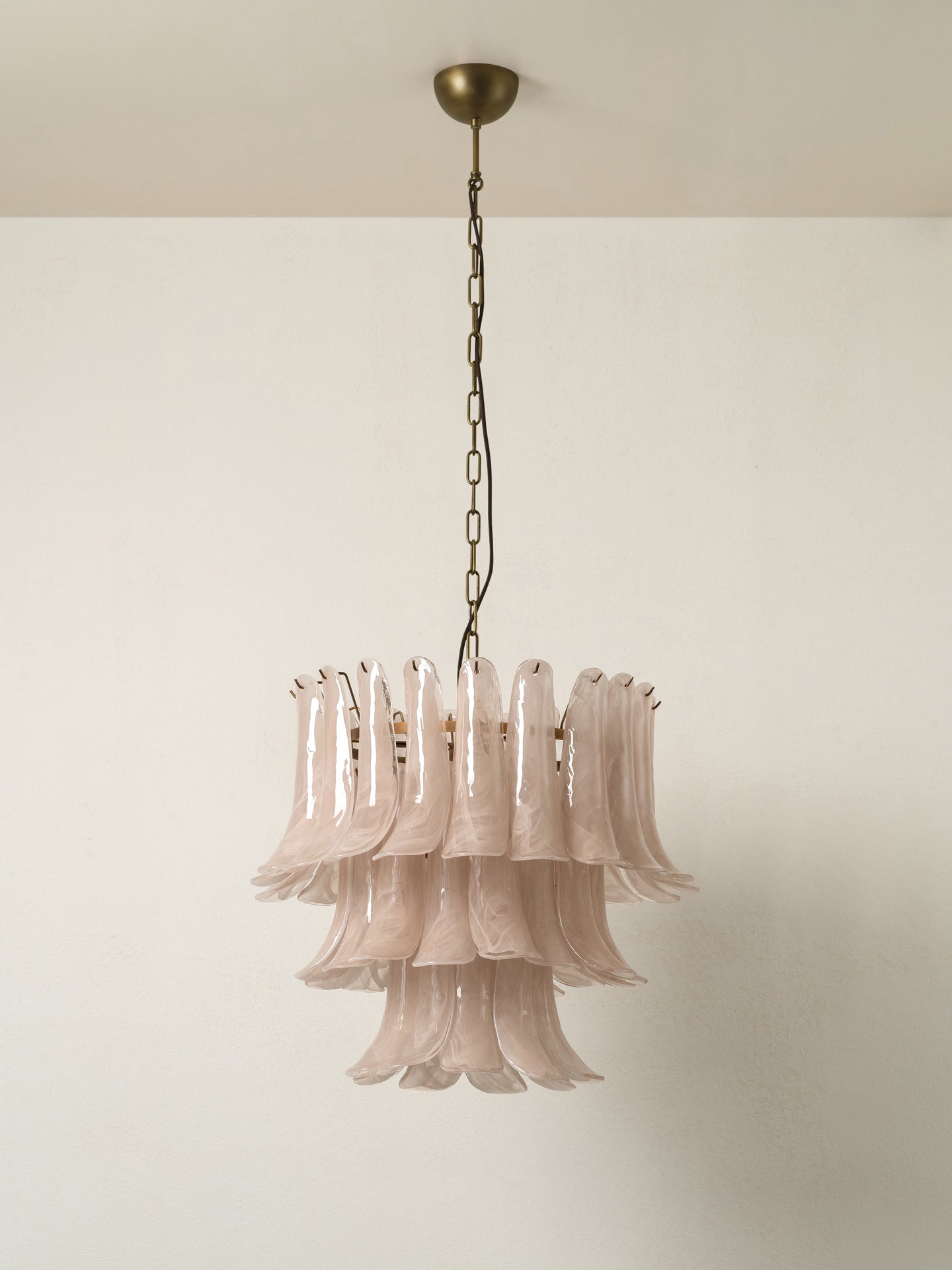 A tiered glass chandelier with fluted, bell-shaped glass shades in a frosted blush pink, suspended from an antique brass chain and ceiling canopy against a plain off-white wall.