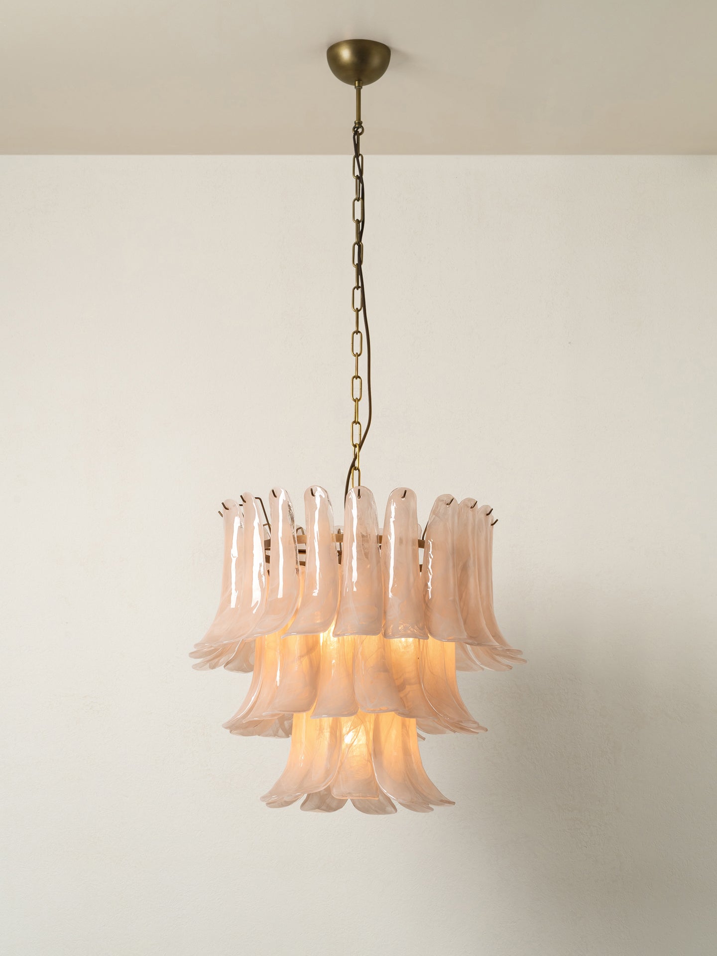 A tiered glass chandelier with flared, bell-shaped translucent shades in a pale blush pink, suspended from a brass chain and ceiling mount against a plain off-white wall.