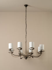 A six-light chandelier with a dark metal leaf-style frame and white cylindrical drum shades, hanging from a chain against a beige wall.