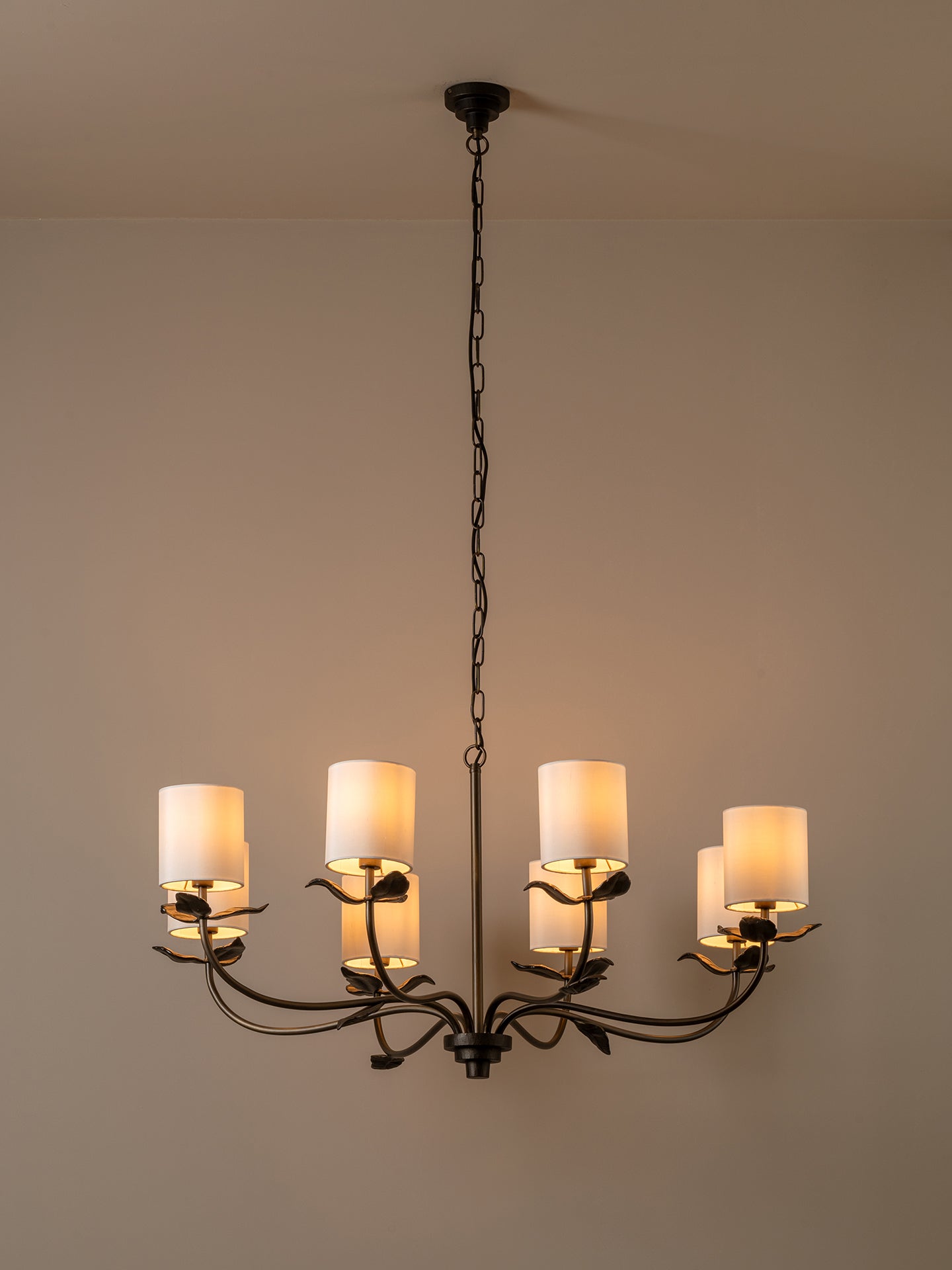 A six-light chandelier with a dark metal frame and warm white cylindrical shades, hanging from a chain against a plain beige wall.