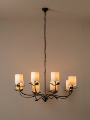 A six-light chandelier with a dark metal frame and warm white cylindrical shades, hanging from a chain against a plain beige wall.