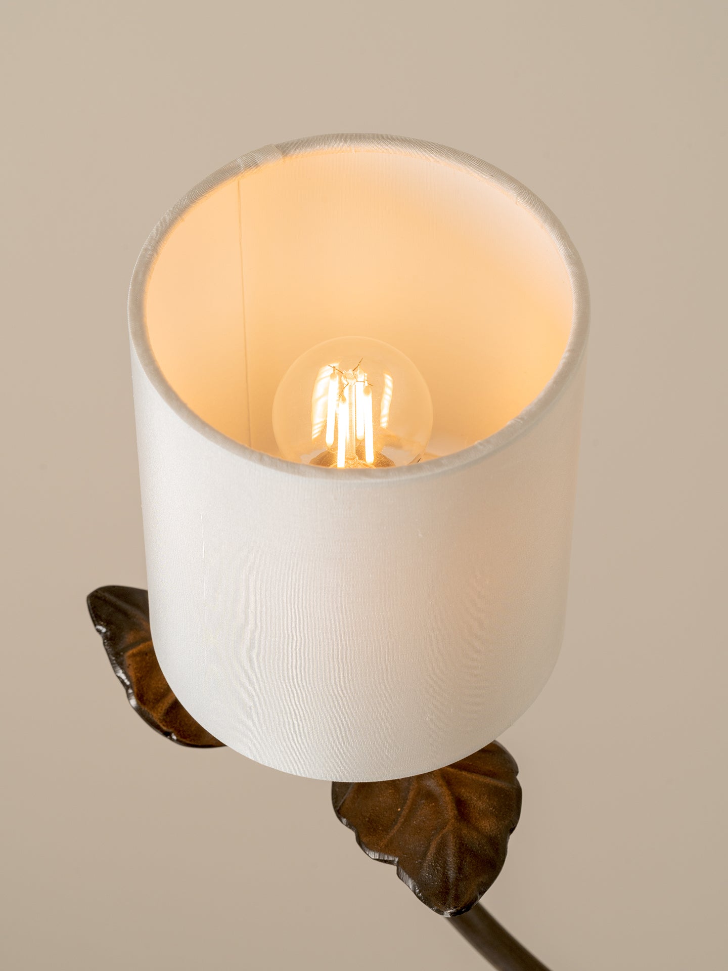 A close-up of a cylindrical white fabric or paper lampshade with a lit Edison-style bulb inside, mounted on a dark metal leaf-shaped wall sconce against a warm beige background.