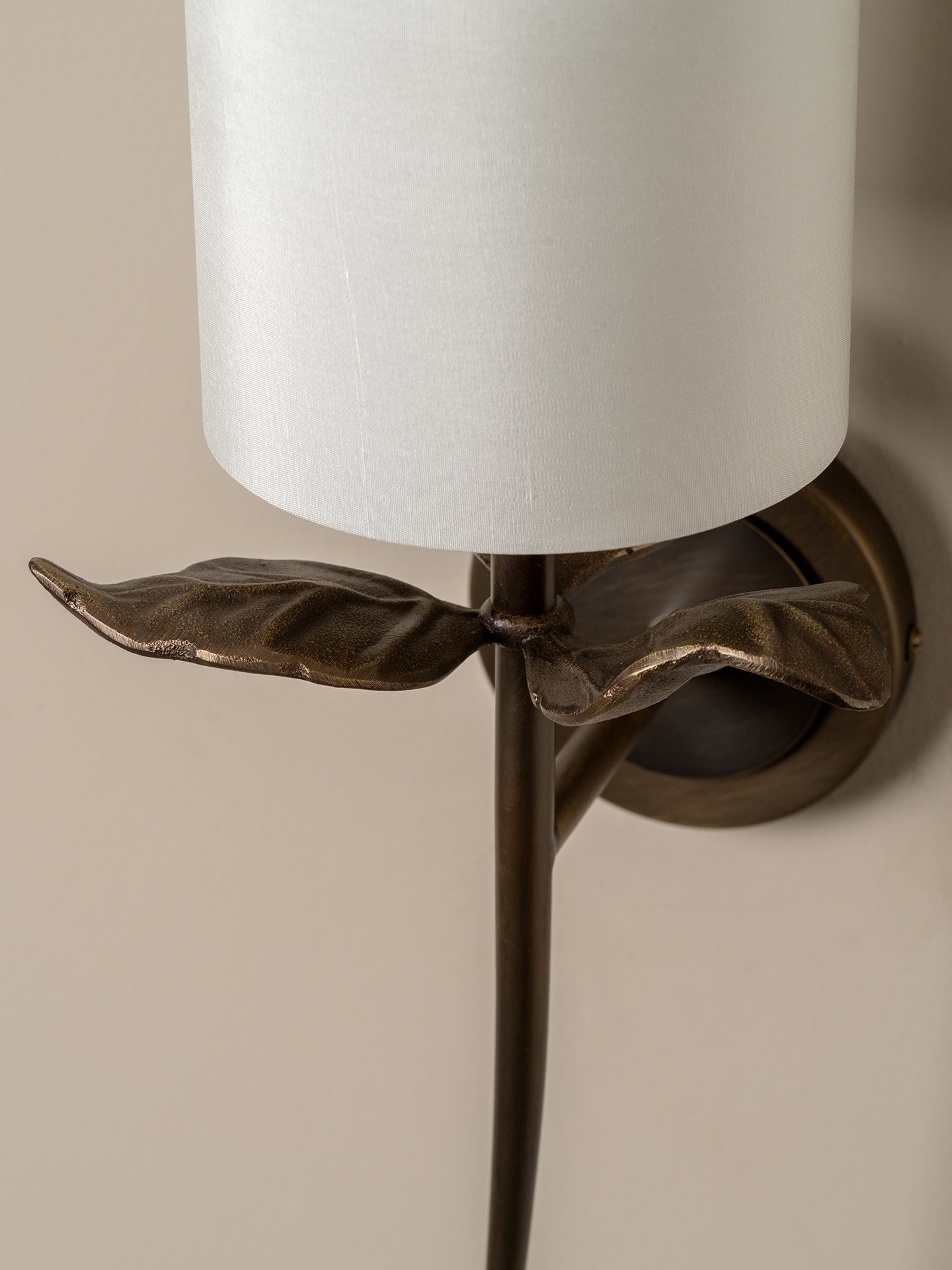 Close-up of a wall-mounted sconce with a white cylindrical drum shade and an ornate bronze metal stem shaped like leaves, mounted on a beige wall.