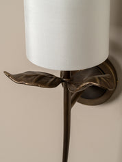 Close-up of a wall-mounted sconce with a white cylindrical drum shade and an ornate bronze metal stem shaped like leaves, mounted on a beige wall.