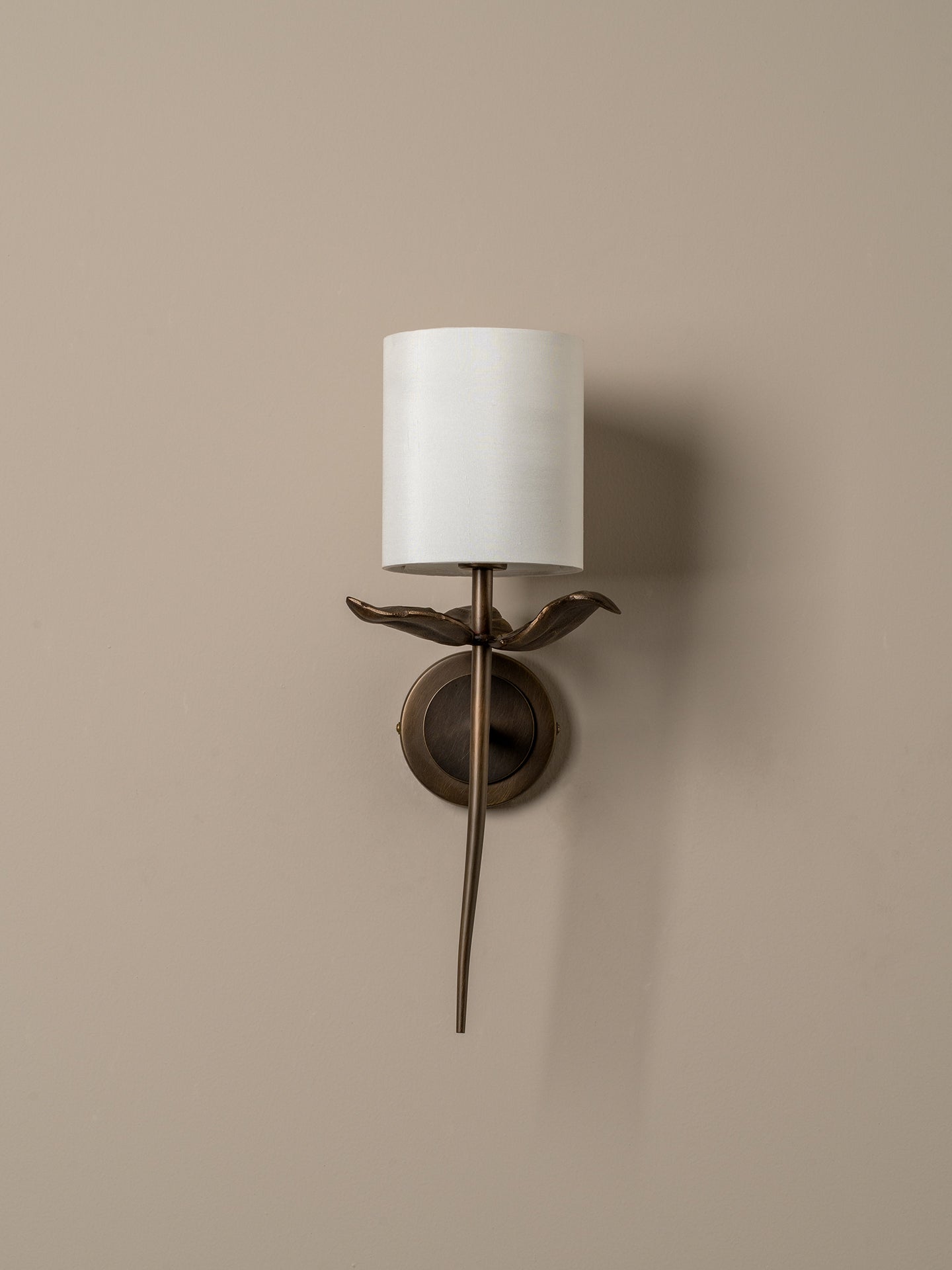 A wall-mounted sconce with a white cylindrical drum shade, a bronze bird-shaped stem, and a circular bronze wall mount, shown against a warm beige wall.