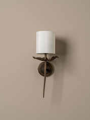 A wall-mounted sconce with a white cylindrical drum shade, a bronze bird-shaped stem, and a circular bronze wall mount, shown against a warm beige wall.