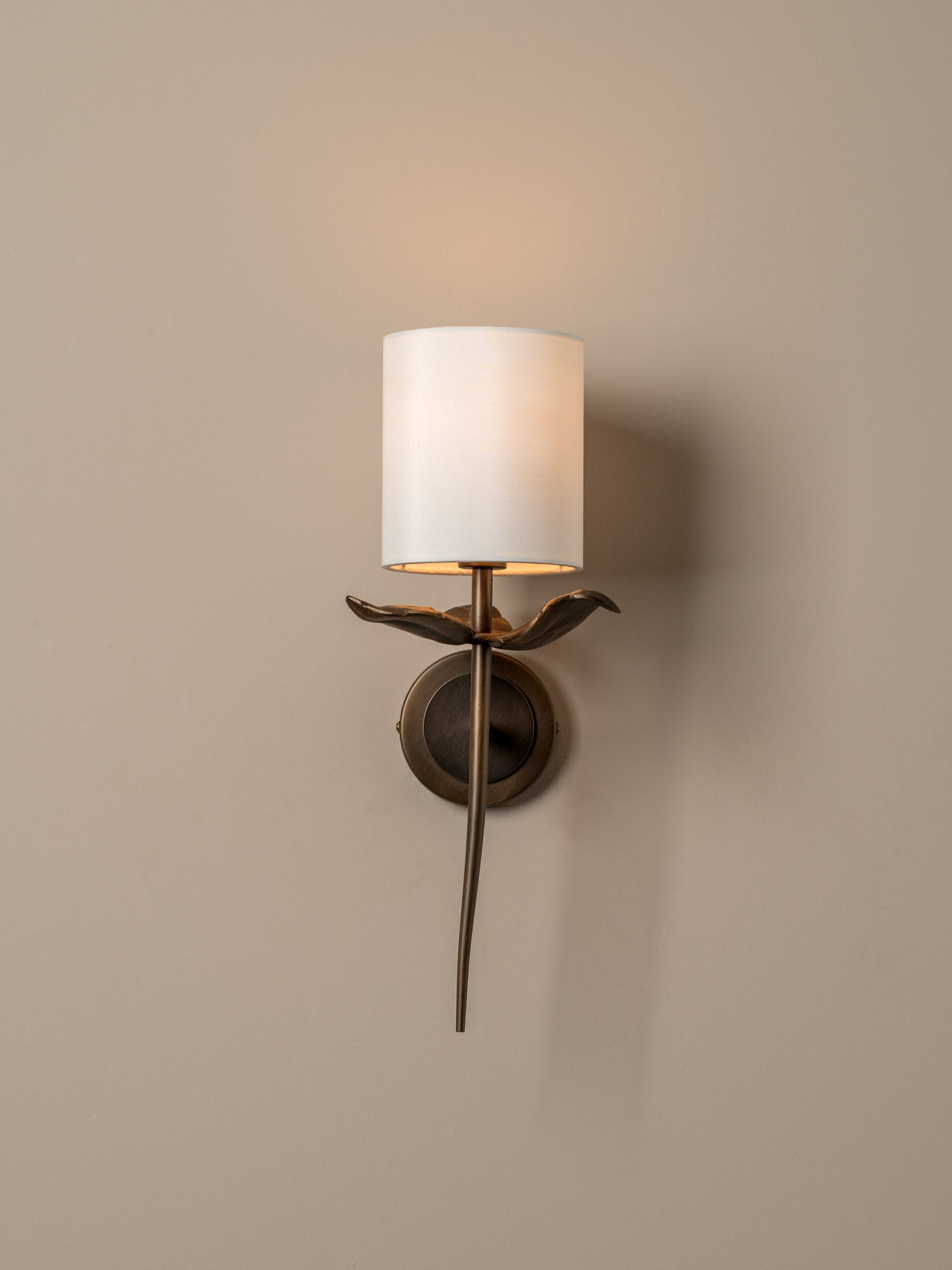 A wall-mounted sconce with a cylindrical white fabric shade, a bronze bird-shaped arm, and a round bronze wall plate, shown lit against a warm beige wall.