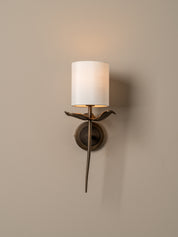 A wall-mounted sconce with a cylindrical white fabric shade, a bronze bird-shaped arm, and a round bronze wall plate, shown lit against a warm beige wall.