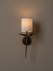 A wall-mounted sconce with a cylindrical white fabric shade, a dark metal bird-shaped arm, and a warm glowing light, set against a plain beige wall.