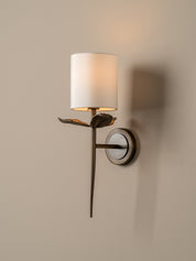 A modern wall sconce with a white cylindrical fabric shade, a dark bronze metal stem shaped like a leaf, and a round wall mount, shown lit against a warm beige wall.