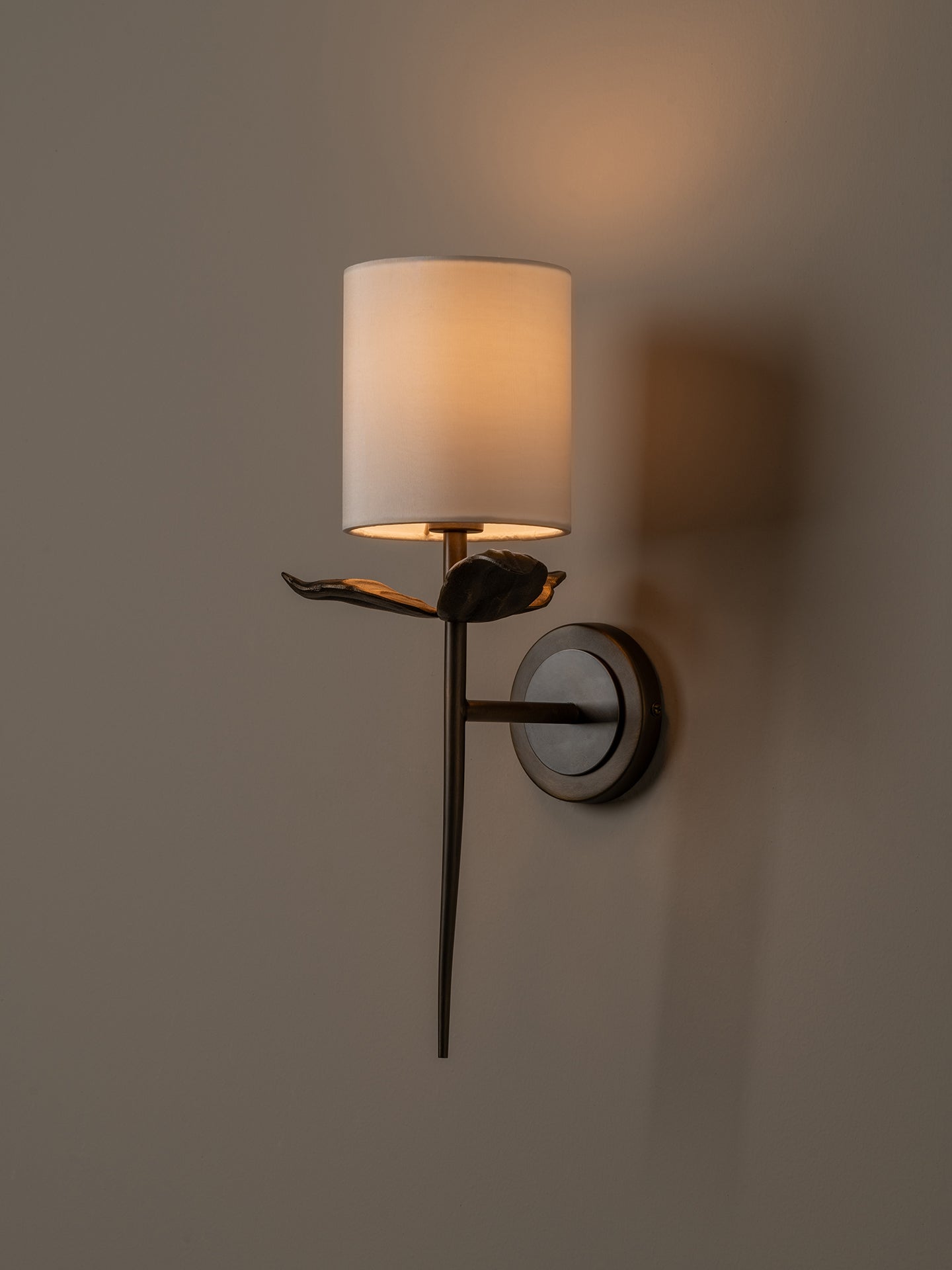 A wall-mounted sconce with a cylindrical white fabric shade, a dark metal branch-like arm, and a circular wall plate, shown lit against a warm neutral wall.