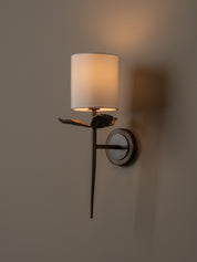 A wall-mounted sconce with a cylindrical white fabric shade, a dark metal branch-like arm, and a circular wall plate, shown lit against a warm neutral wall.