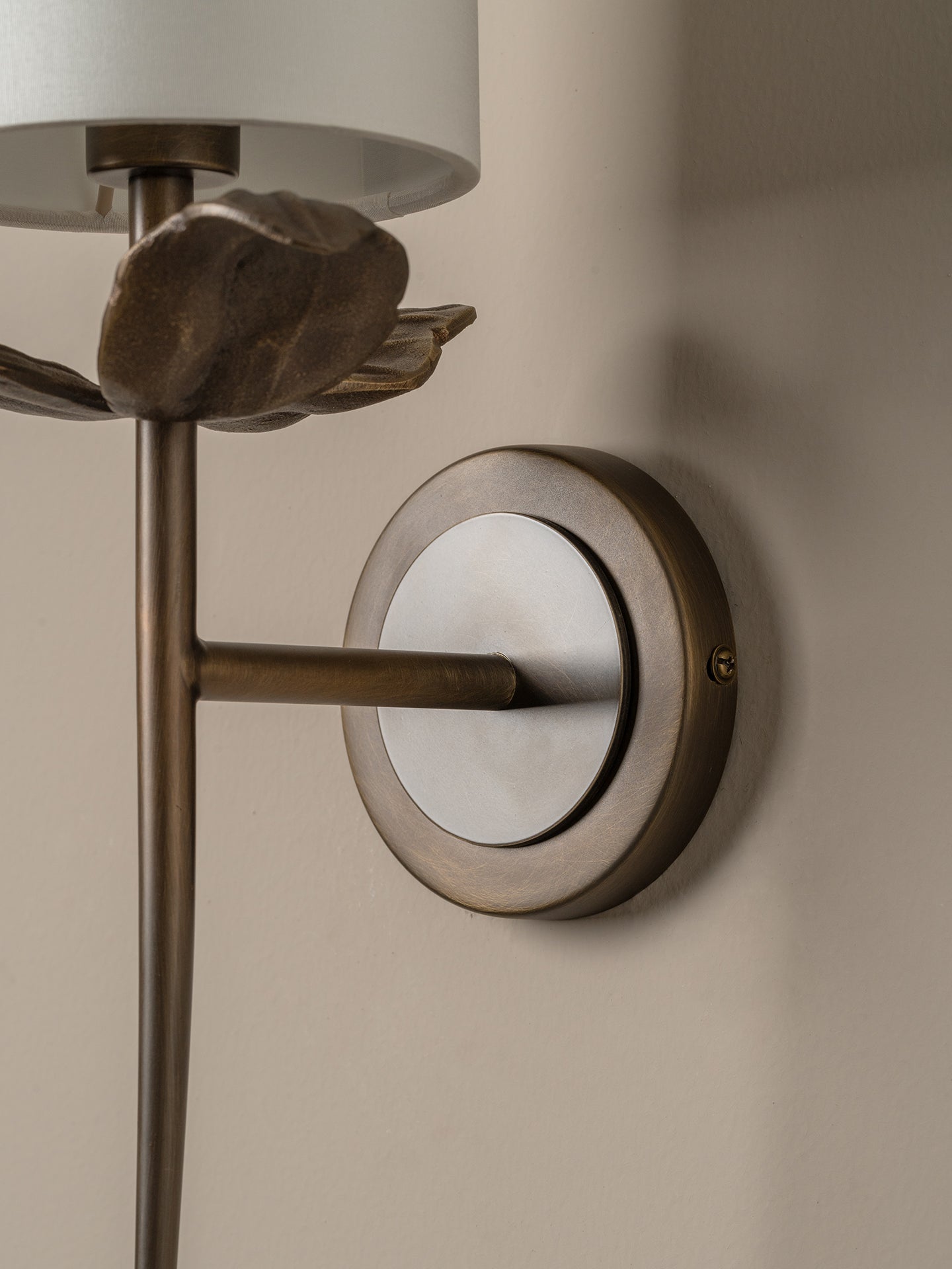 Close-up of a wall-mounted sconce with a brushed metal circular backplate, vertical stem, sculptural leaf detail, and part of a white drum shade against a warm beige wall.