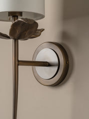 Close-up of a wall-mounted sconce with a brushed metal circular backplate, vertical stem, sculptural leaf detail, and part of a white drum shade against a warm beige wall.