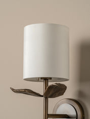 A close-up of a wall-mounted sconce with a white cylindrical drum shade, a bronze leaf-shaped decorative element, and a matching round wall mount against a warm beige wall.