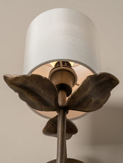 Close-up of a decorative wall sconce with a white fabric drum shade, bronze metal stem, and sculptural leaf-like side details against a warm beige wall.