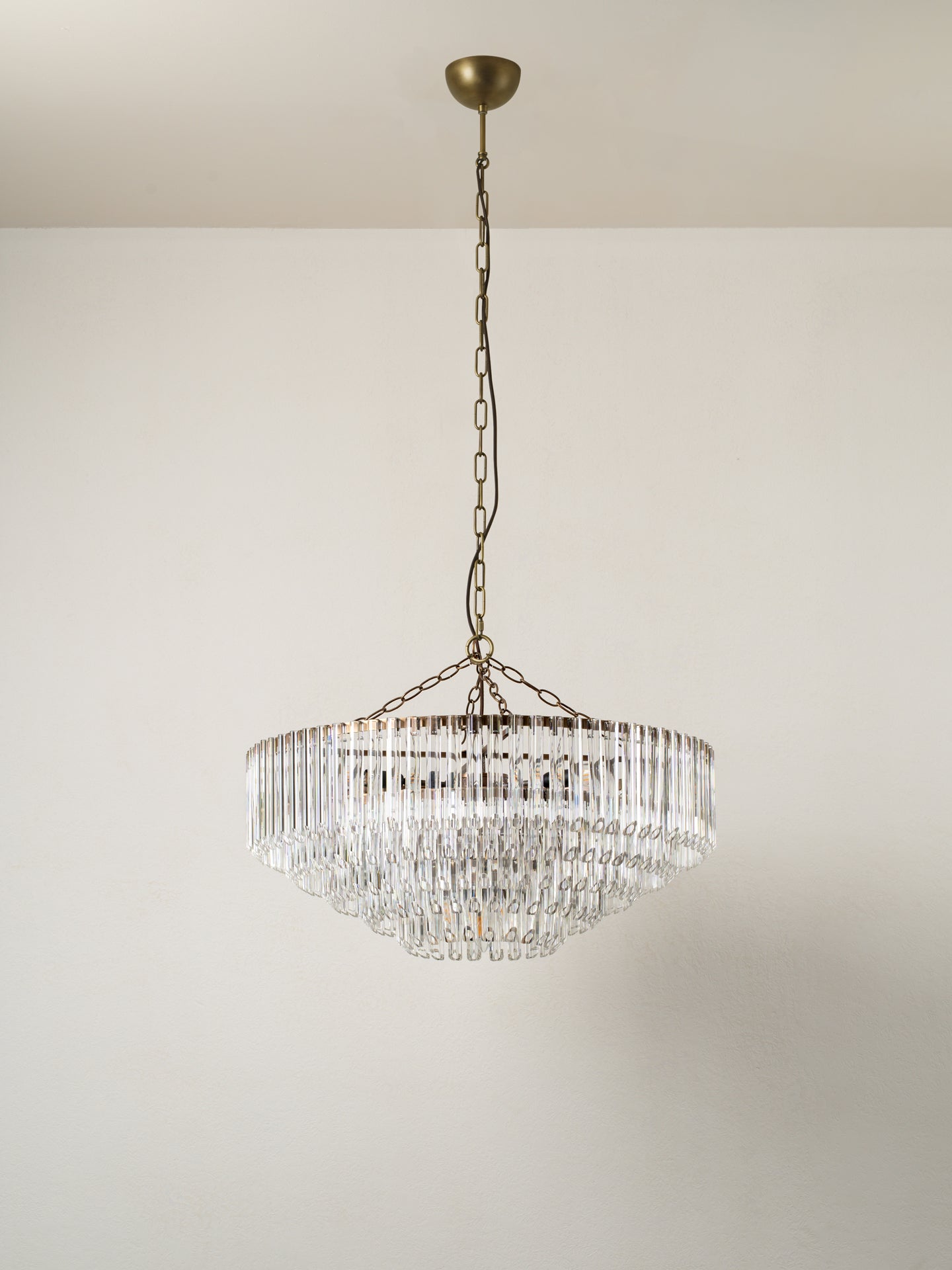 A tiered crystal chandelier with an antique bronze metal frame and chain, suspended from a ceiling against a plain off-white wall.