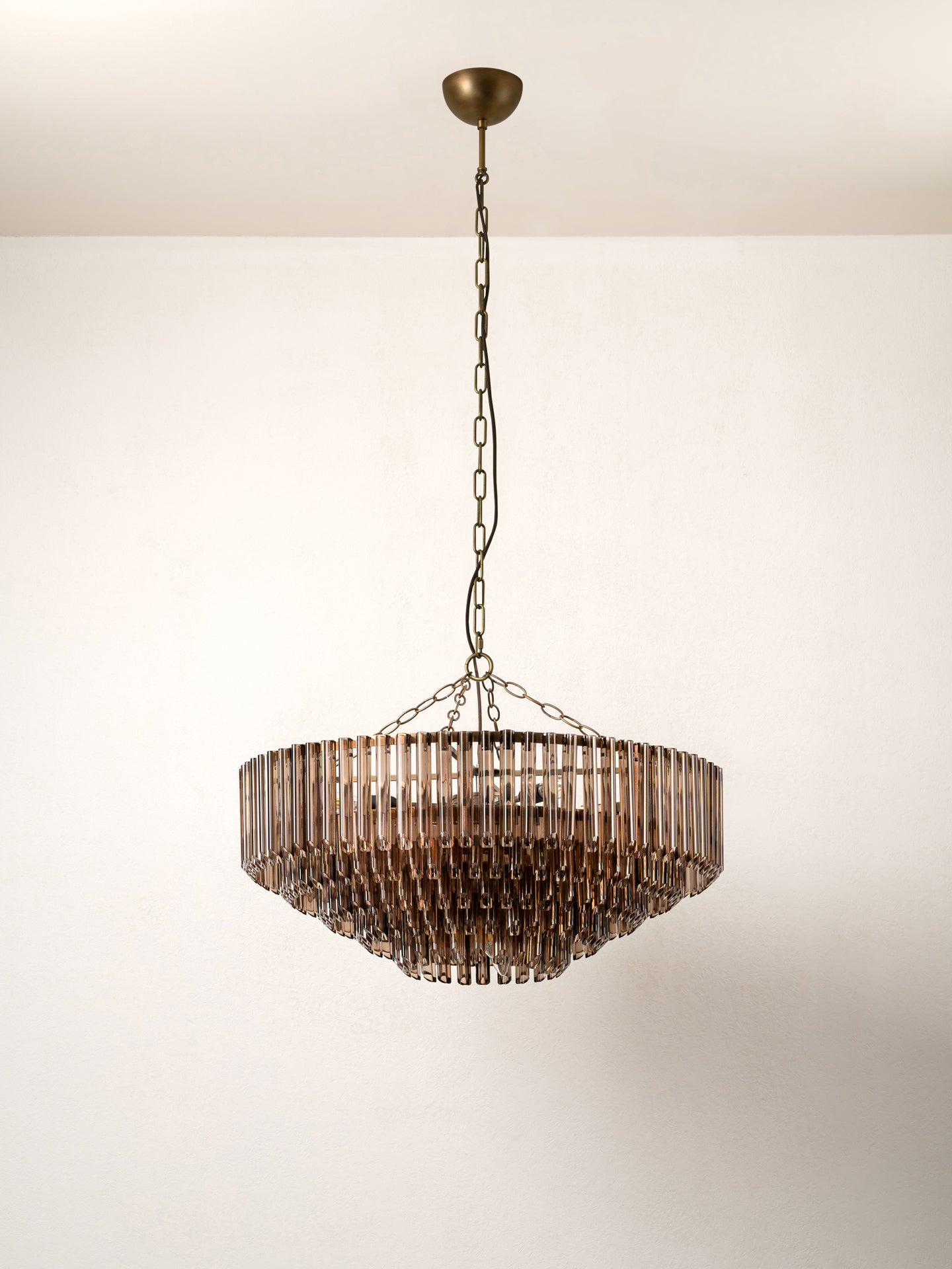 A round pendant chandelier with a bronze-finished metal frame and hanging glass shades, suspended from a chain against a plain off-white wall.