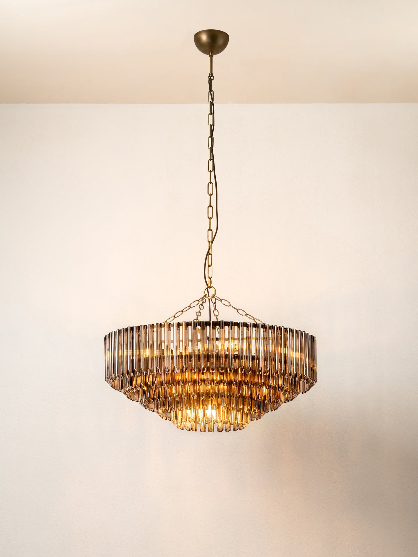 A modern tiered chandelier with a bronze-finished metal frame and hanging chain, featuring clear ribbed glass shades that glow warm yellow when lit, shown against a plain off-white wall and ceiling.