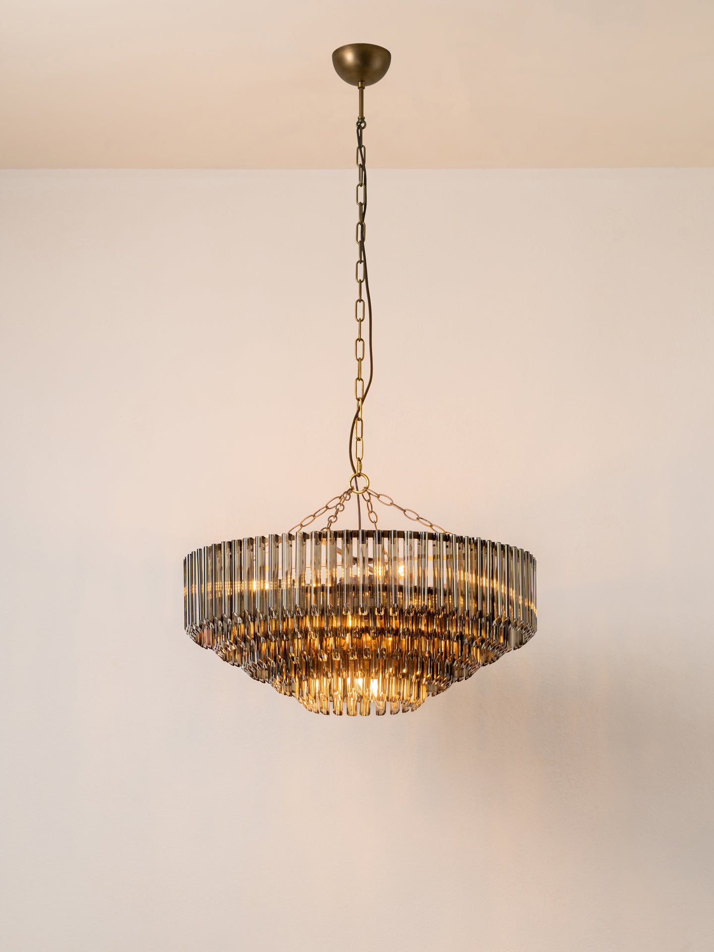 A tiered chandelier with a bronze metal frame and clear glass cylindrical prisms, suspended by a chain from a matching ceiling canopy against a plain off-white wall.