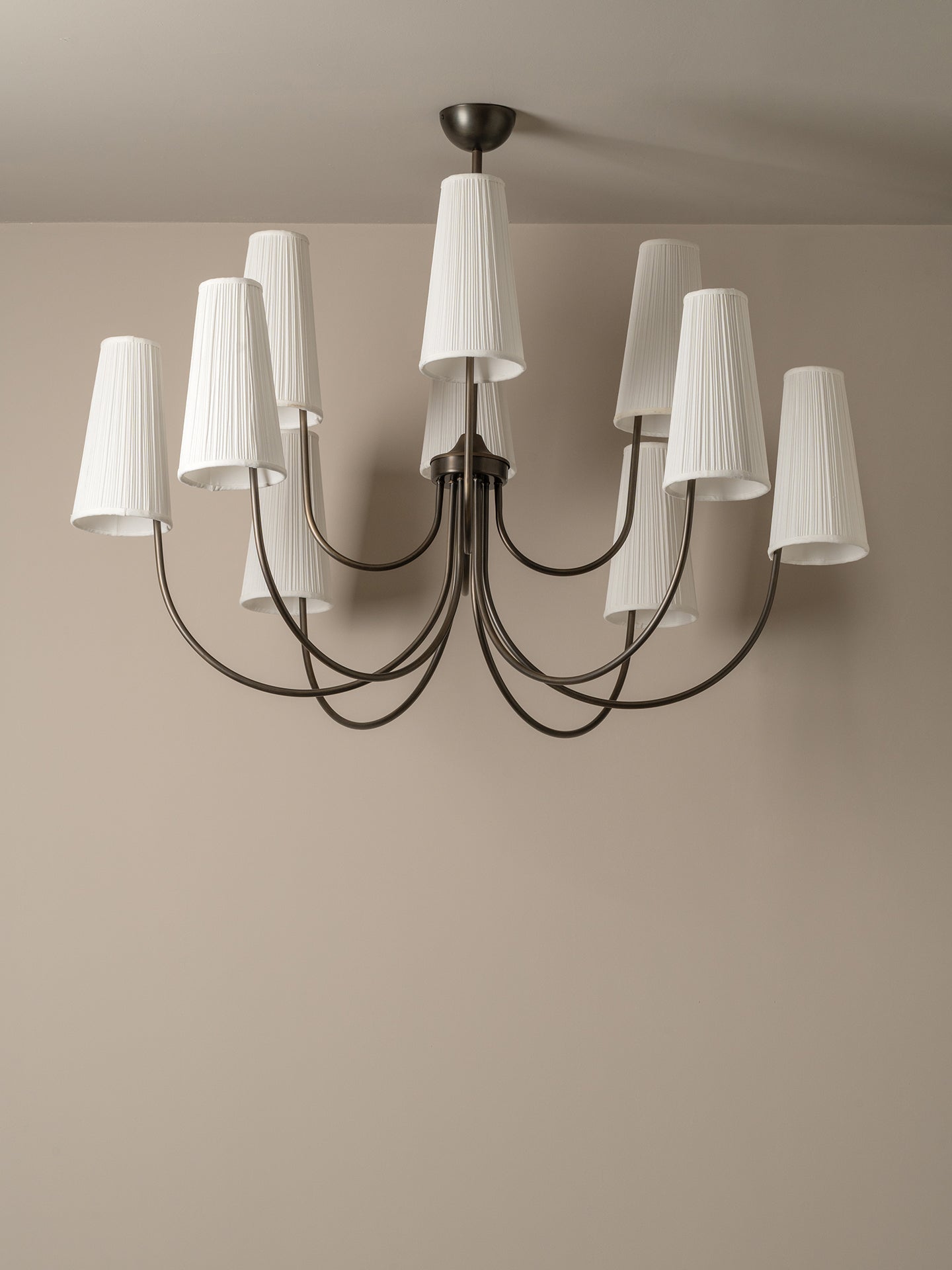 A modern multi-arm chandelier with a dark metal frame and white pleated drum shades, shown hanging against a neutral beige wall and ceiling.