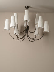 A modern multi-arm chandelier with a dark metal frame and white pleated drum shades, shown hanging against a neutral beige wall and ceiling.