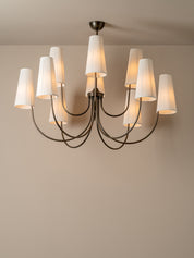 A modern multi-arm chandelier with white pleated drum shades and a dark metal frame, shown lit against a neutral beige wall and ceiling.