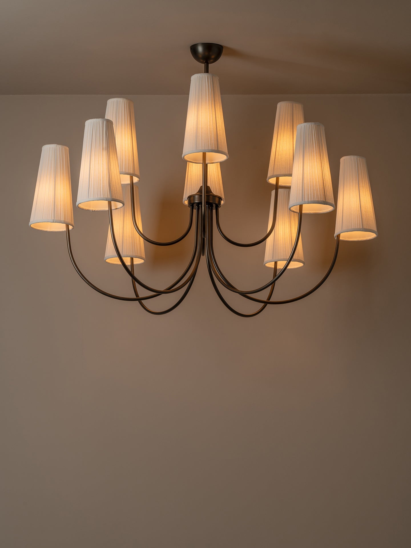 A multi-light chandelier with a dark metal frame and nine tapered white fabric shades, glowing with warm light against a neutral beige wall and ceiling.