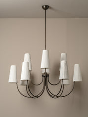 A modern nine-light chandelier with a dark metal frame and pleated white drum shades, shown hanging against a neutral beige wall.