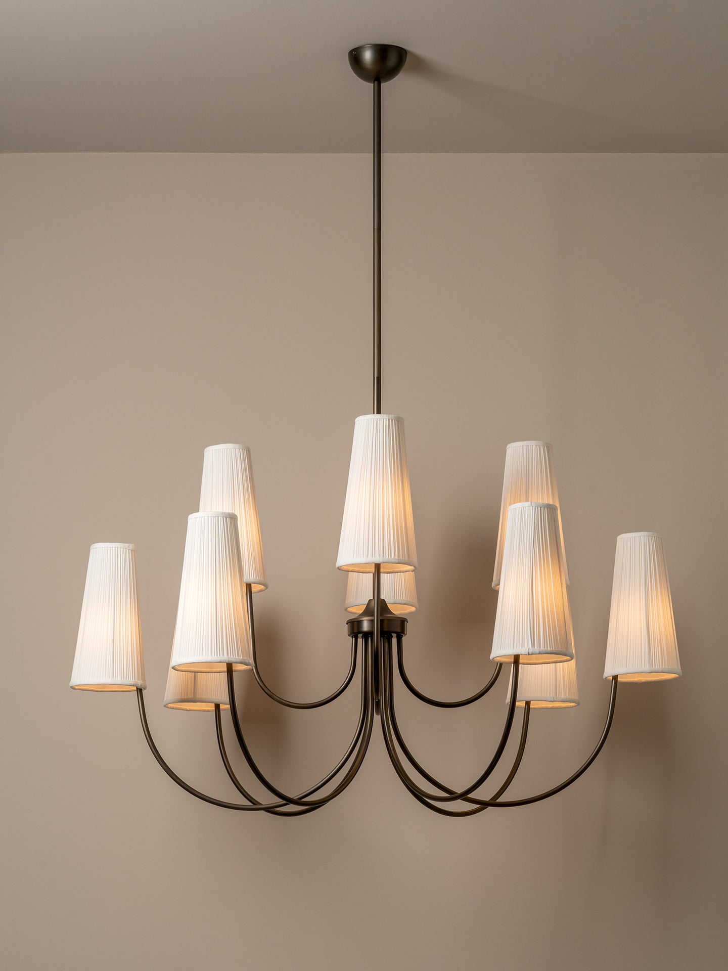 A modern multi-light chandelier with a dark metal frame and eight white pleated drum shades, shown illuminated against a neutral beige wall.