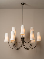 A modern multi-light chandelier with a dark metal frame and eight white pleated drum shades, shown illuminated against a neutral beige wall.