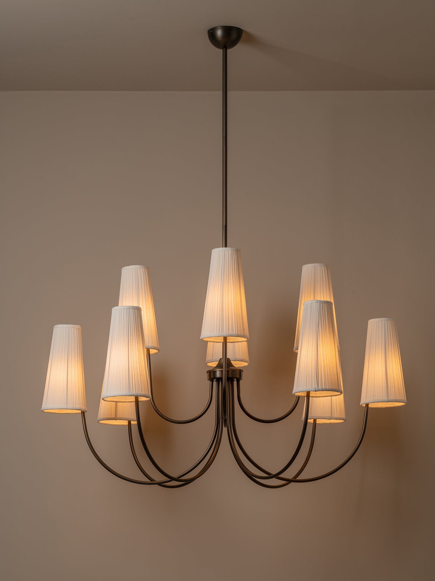 A modern multi-light chandelier with a dark metal frame and eight tapered white fabric shades, shown illuminated against a plain beige wall.
