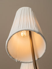 A close-up of a pleated white fabric lampshade with a warm glowing light bulb inside, mounted on a metallic gold stand against a neutral beige background.