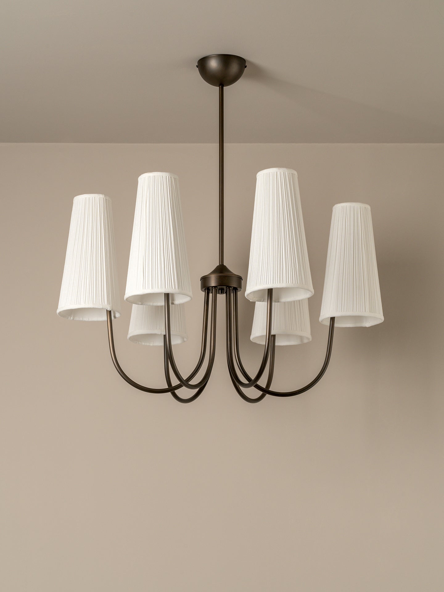 A six-light chandelier with a dark bronze metal frame and white pleated drum shades, shown hanging from a ceiling against a beige wall.