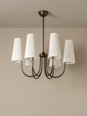 A six-light chandelier with a dark bronze metal frame and white pleated drum shades, shown hanging from a ceiling against a beige wall.